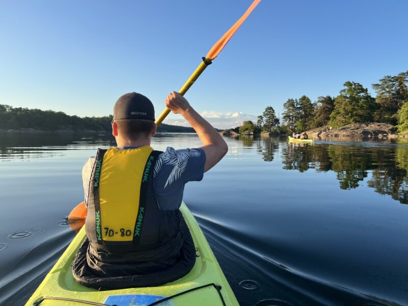 Person som paddlar kajak på lugna vatten med omgivande träd och klar himmel i bakgrunden. En annan grupp människor ses paddla kajak i fjärran.